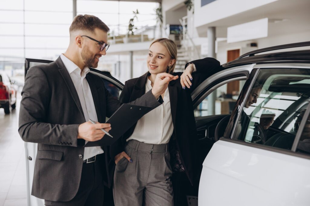 Car Sales Manager Showing Auto To Caucasian Lady Buyer Standing In Luxury Automobile Dealership