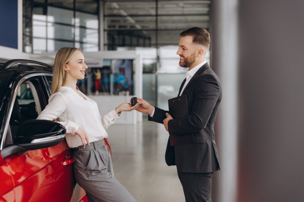 Car Sales Manager Showing Auto To Caucasian Lady Buyer Standing In Luxury Automobile Dealership
